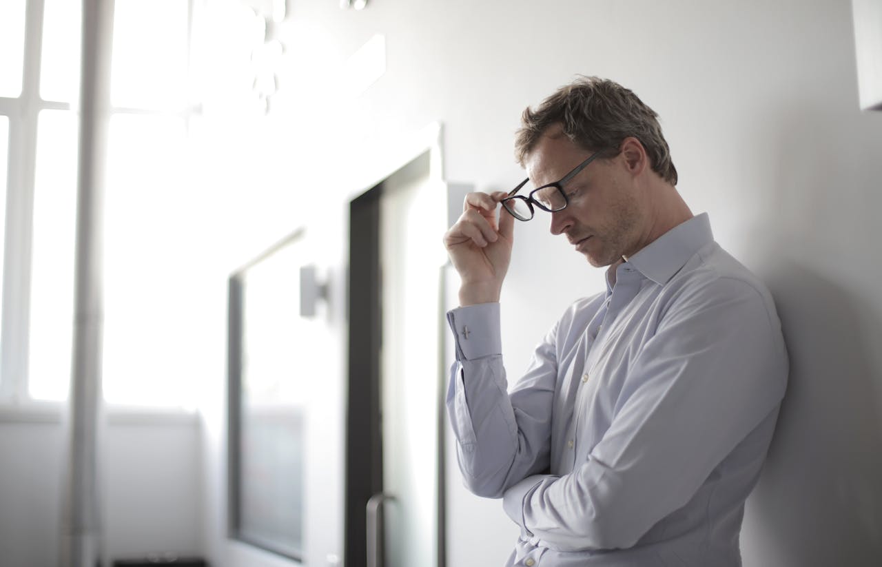 gallery-4 Thoughtful man in a bright room holding his glasses while leaning against a wall.