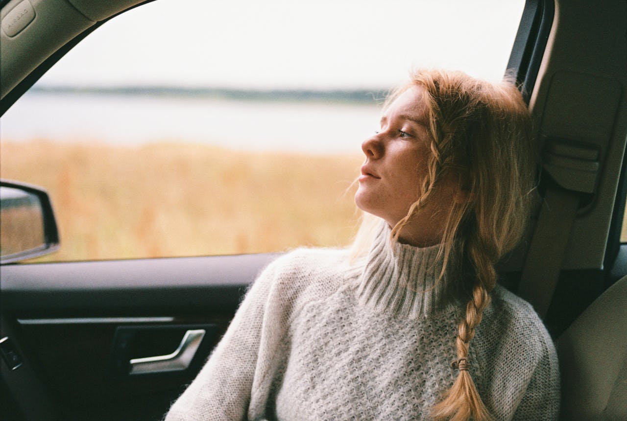 hero-img-01 A thoughtful woman with braided hair gazes out a car window during a daytime road trip.